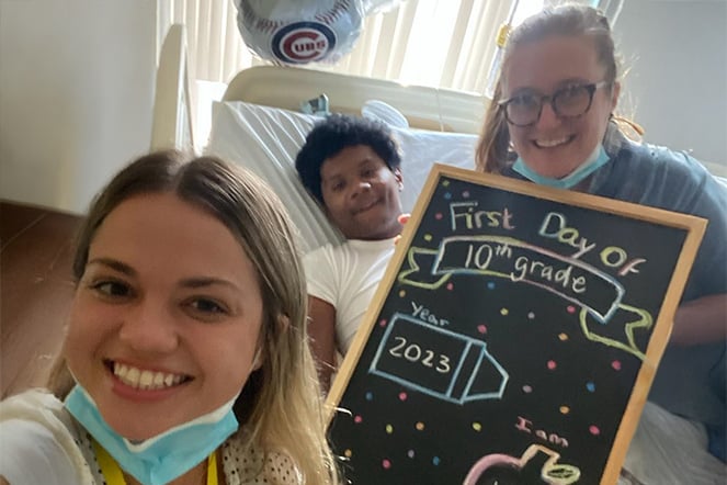 an adult takes a selfie photo with a child in a hospital bed and an adult holding a chalkboard sign that reads "First Day of 10th Grade 2023"