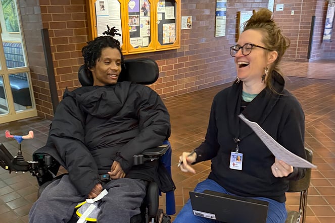 a child in a power chair laughs next to an adult holding a stack of papers