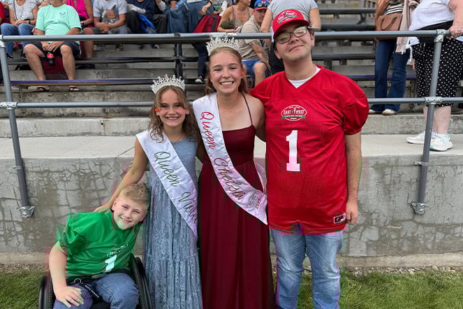four children in front of the bleachers at a football game. The two children in the middle are wearing gowns, tiaras, and sashes. The two children on the ends are wearing football jerseys