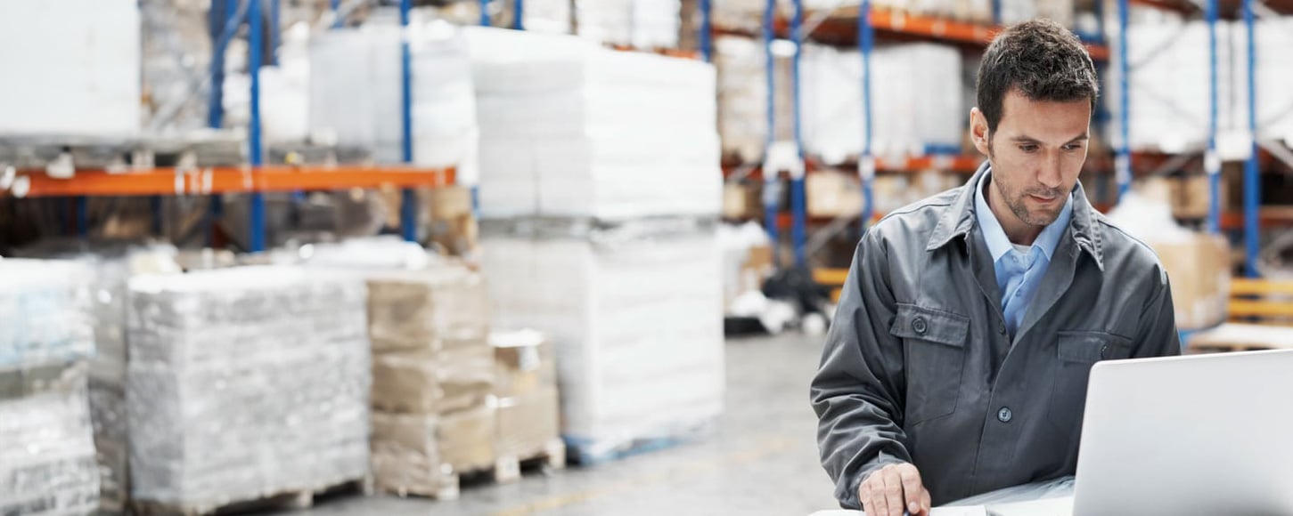 A young man using his laptop and checking his notes while working in a warehouse
