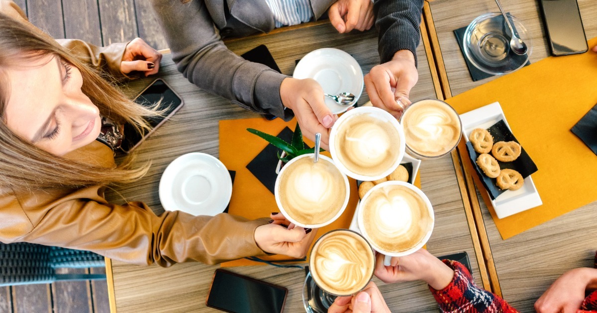 Group raising cups of latte art together over a table with pastries and phones.