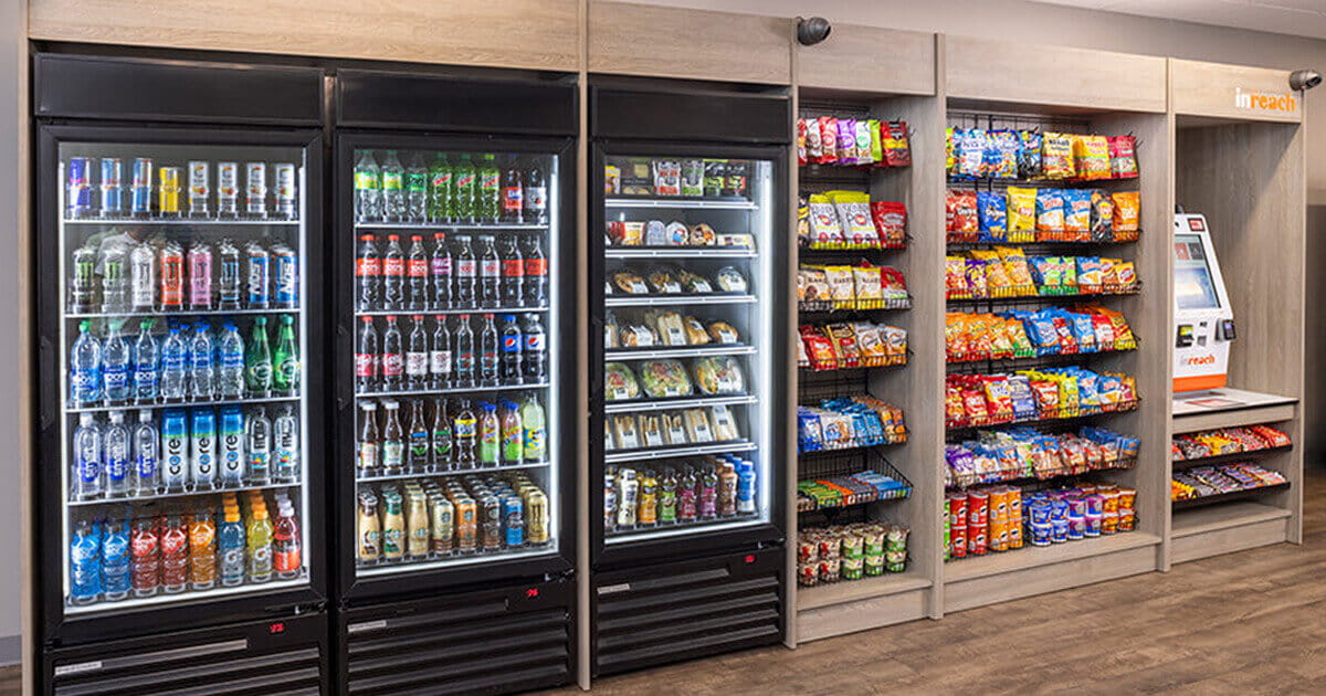 Self-service micro market with refrigerated coolers stocked with drinks and packaged meals, shelves filled with assorted snacks, and a payment kiosk in a modern break room setting.