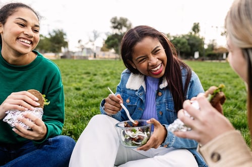 A group of college students eating a salad and sitting on a lawn.