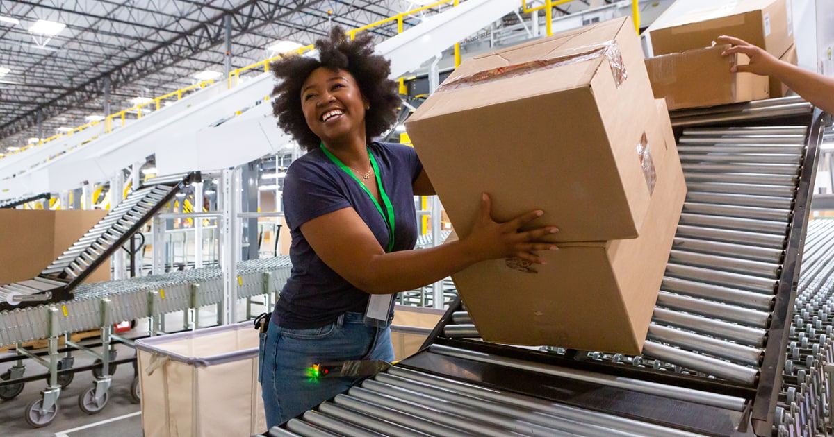 Worker lifting a large cardboard box onto a conveyor belt inside a busy warehouse with automated sorting equipment.