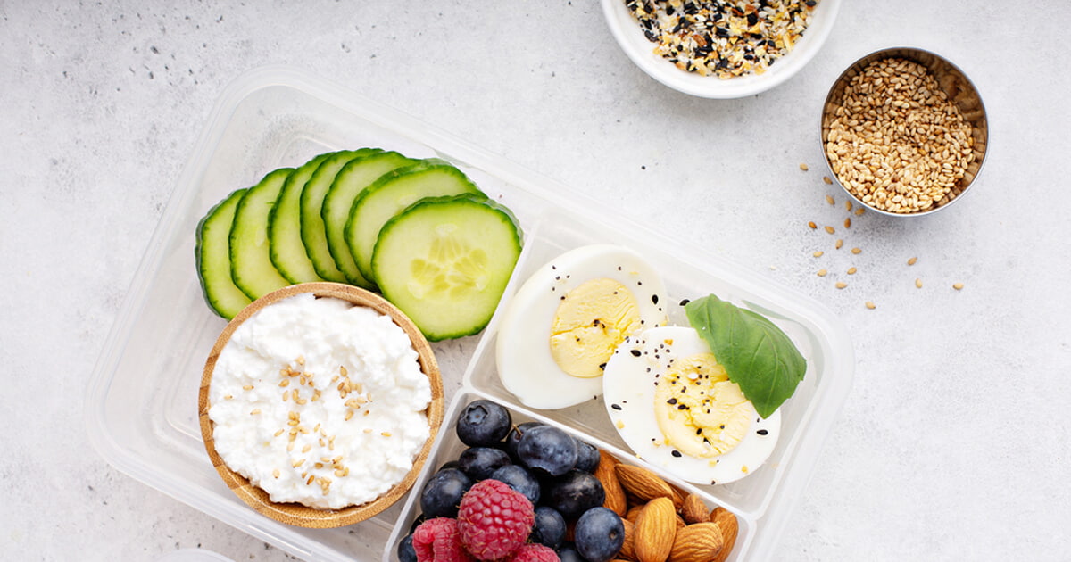 Meal prep container with sliced cucumber, cottage cheese, hard-boiled eggs, fresh blueberries, raspberries, and almonds, with a small bowl of seeds on the side.