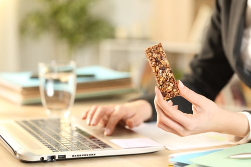 A person holding a granola bar while working on a laptop.