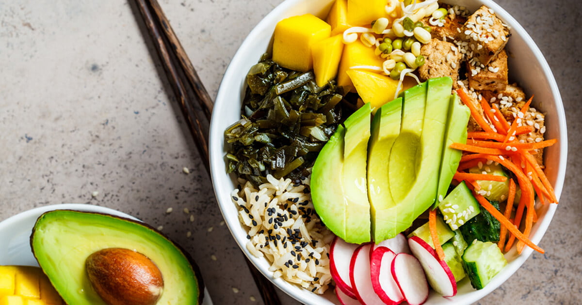 Colorful poke bowl with sliced avocado, mango cubes, radish, carrots, cucumber, tofu, seaweed, and rice topped with sesame seeds, served with half an avocado on the side.