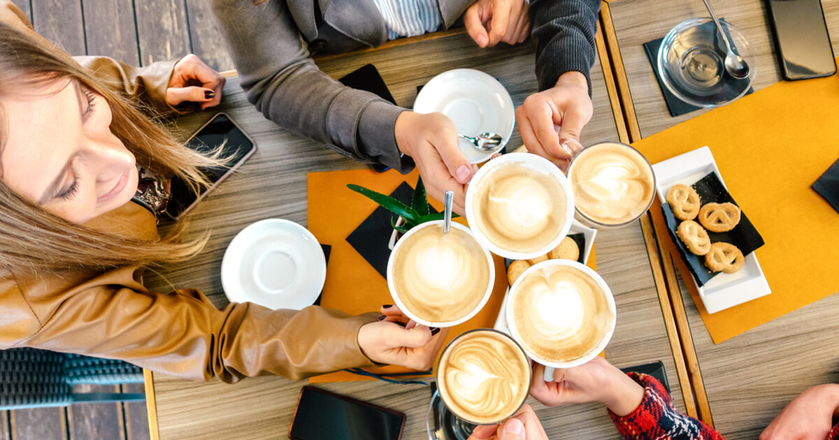 Group raising cups of latte art together over a table with pastries and phones.