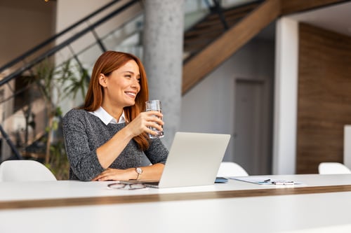 A woman drinking a glass of water while working on a laptop.
