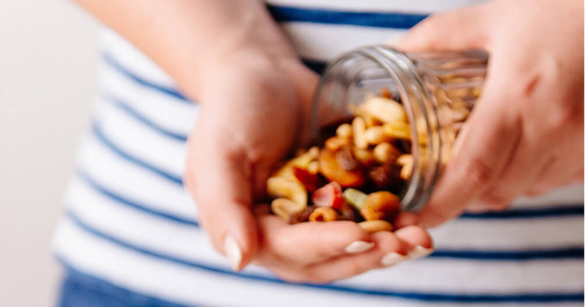 Hands sprinkling mixed nuts from a small jar over a bowl of oatmeal topped with sliced apples on a white table.