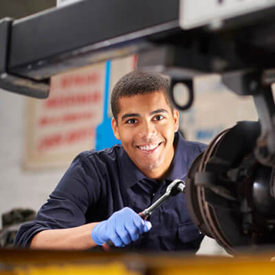 Mechanic wearing gloves uses a wrench to work on a vehicle part in an auto repair shop, with tools and equipment visible in the background.