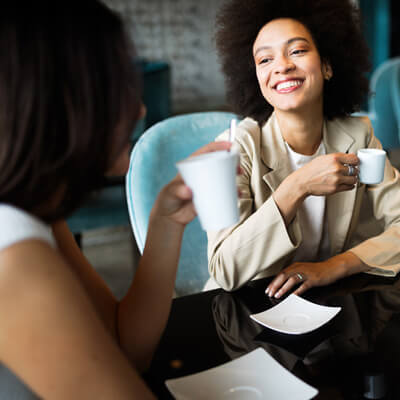 Two people sit at a black table in a modern café, holding white coffee cups with empty white plates in front of them, teal chairs and a soft gray wall in the background.