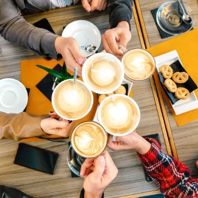 Group raising cups of latte art together over a table with pastries and phones.
