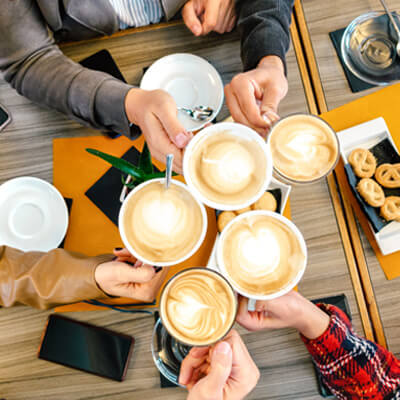 Group raising cups of latte art together over a table with pastries and phones.