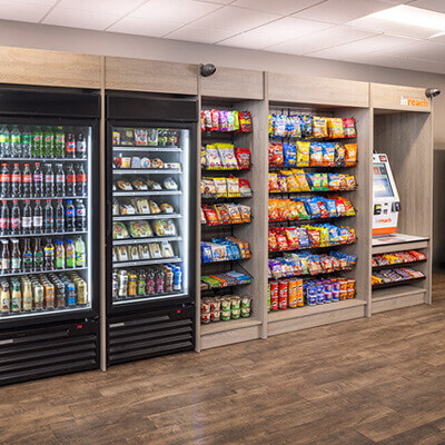 Self-service micro market with refrigerated coolers stocked with drinks and packaged meals, shelves filled with assorted snacks, and a payment kiosk in a modern break room setting.