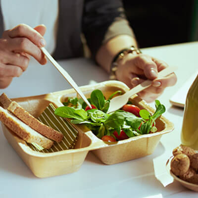 Person eating a fresh salad with cherry tomatoes and spinach from a compostable container, with slices of bread and a drink nearby.