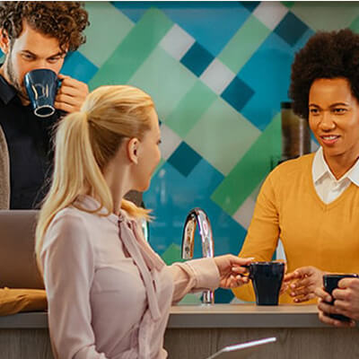Group of people gathered in a break area, chatting and holding coffee near a counter with a colorful backdrop.