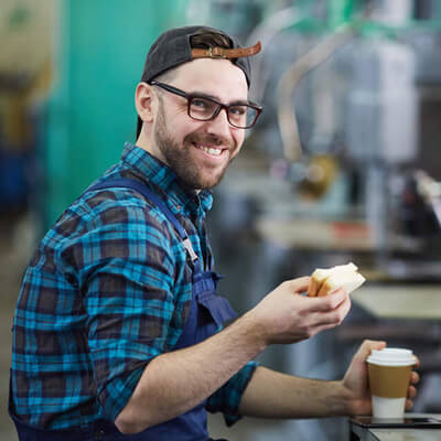 Worker in a plaid shirt and apron taking a break while holding a sandwich and a cup of coffee in an industrial setting.