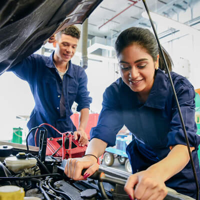 Two mechanics in blue coveralls working under an open car hood in a brightly lit workshop with tools and equipment visible.