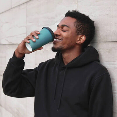 Person in a black hoodie drinking from a teal takeaway cup while leaning against a stone wall.
