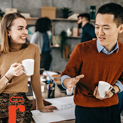 Coworkers standing and chatting in an office while holding coffee cups.