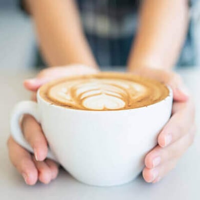 Hands holding a white ceramic cup filled with latte featuring heart-shaped foam art on top, placed on a light-colored surface.