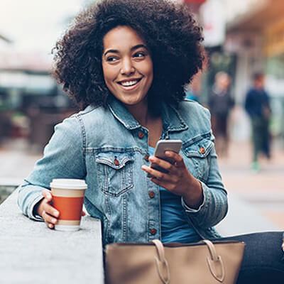 Person sitting outdoors holding a smartphone in one hand and a takeaway coffee cup in the other, with a beige handbag on the bench nearby.
