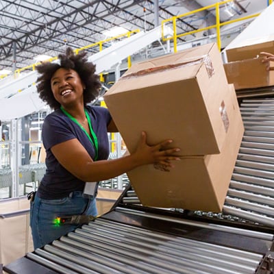 Worker lifting a large cardboard box onto a conveyor belt inside a busy warehouse with automated sorting equipment.