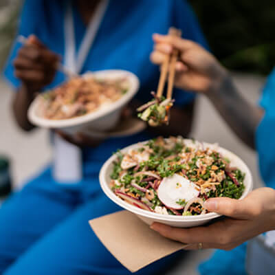 People in blue scrubs eating fresh salad bowls with chopsticks during a meal break.