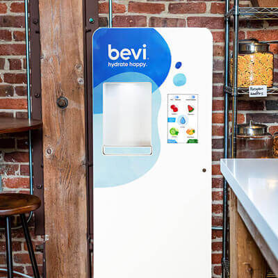 Bevi water dispenser with a white and blue design placed against an exposed brick wall, next to wooden beams and metal shelving with jars in a modern kitchen setting.
