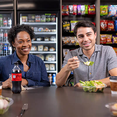 People eating and chatting at a table in a break area with coolers and snack shelves in the background.