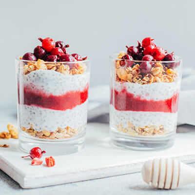 Two glasses filled with layered chia pudding, granola, and berry puree, topped with fresh red berries on a white surface.
