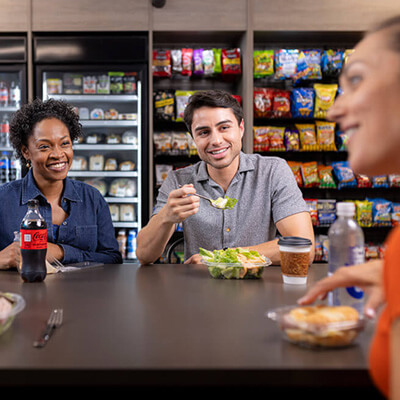 People sitting at a table in a break area with shelves of snacks and drinks in the background, eating salads and drinking beverages.