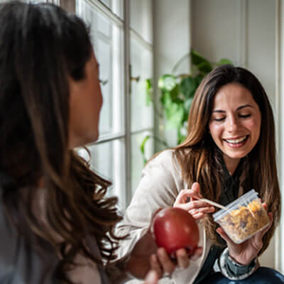 Two people sharing snacks, one holding an apple and the other holding a container of granola.
