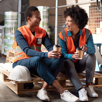 Two workers in orange safety vests sitting on pallets in a warehouse, holding coffee cups during a break.