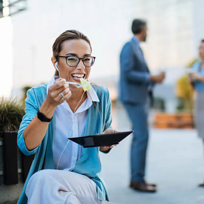 Woman sitting outdoors eating while holding a tablet, with two people conversing in the background.