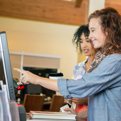 Person pointing at a touchscreen kiosk while another stands nearby in a modern indoor setting.