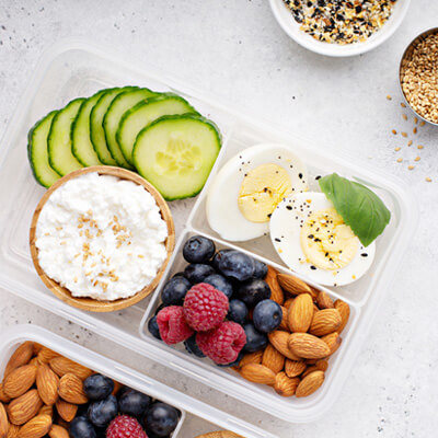 Meal prep container with sliced cucumber, cottage cheese, hard-boiled eggs, fresh blueberries, raspberries, and almonds, with a small bowl of seeds on the side.