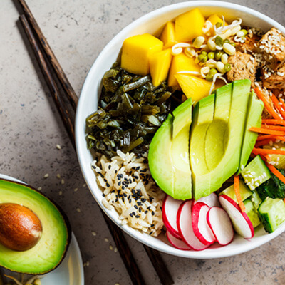 Colorful poke bowl with sliced avocado, mango cubes, radish, carrots, cucumber, tofu, seaweed, and rice topped with sesame seeds, served with half an avocado on the side.