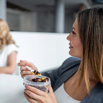Woman eating a container of mixed nuts and dried fruit while another person holds an apple at a desk.