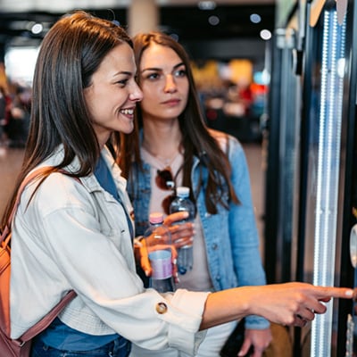 Two people selecting items from a vending machine while holding bottled drinks in a busy indoor setting.