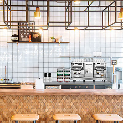 Modern coffee bar with white tiled wall, espresso machines, wooden counter with hexagon pattern, and three stools under geometric light fixtures.
