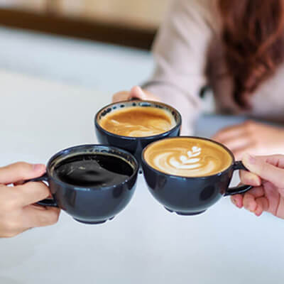 Three people clinking black coffee mugs filled with different coffee drinks over a white table.