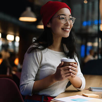 Person holding a takeaway coffee cup while sitting in a cozy café with warm lighting and plants.