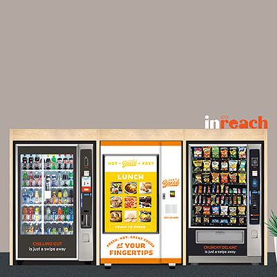 Row of vending machines offering drinks, snacks, and hot meals, with an informational banner on the left and a potted plant on the right against a neutral wall.