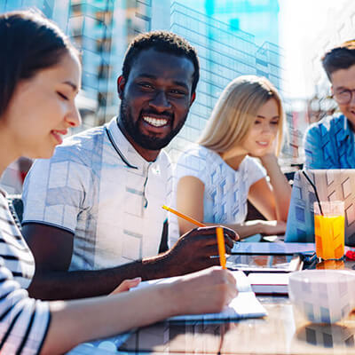 Group of people sitting outdoors at a table working together with notebooks, laptops, and drinks.