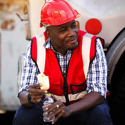 Construction worker in a red safety vest and hard hat taking a break while eating and holding a water bottle.