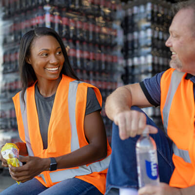 Workers in orange safety vests taking a break with bottled water and a snack in a warehouse setting.