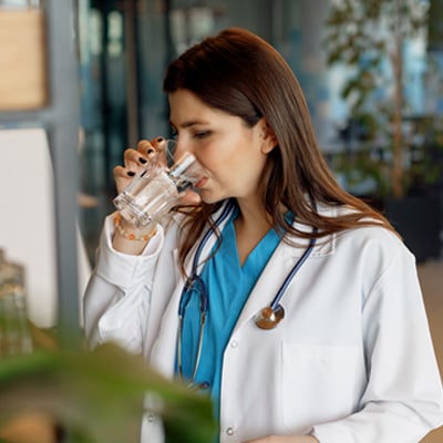 Healthcare worker in scrubs and a white coat drinking water from a clear glass in a modern office setting.