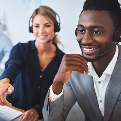 Two people wearing headsets working together, with one person pointing at a screen in a collaborative office setting.